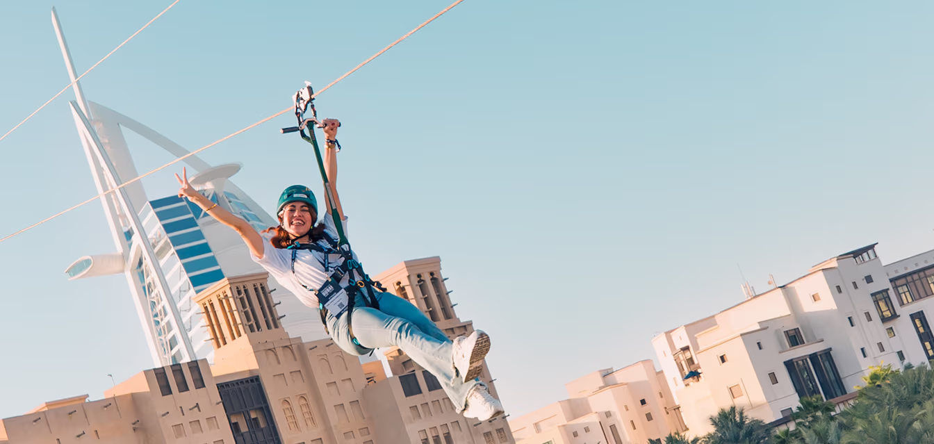 Woman smiling and making a peace sign while zip-lining with Burj Al Arab and city buildings in the background.